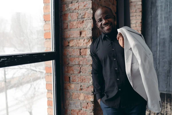 Bonito e elegante homem afro-americano de aparência modelo mantém uma jaqueta em seu ombro e posa em um estúdio loft com grandes janelas — Fotografia de Stock