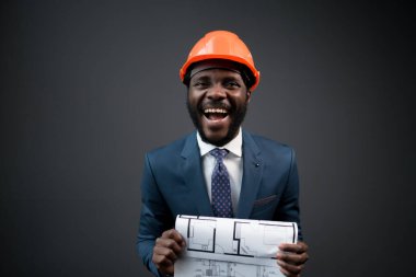 Kameraya gülümseyen Afrikalı Amerikalı bir adam karanlık bir arka planda duruyor ve elinde bir çizim olan bir kağıt parçası tutuyor.