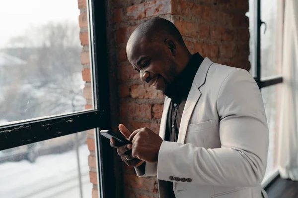 Африканский американец в черной рубашке стоял у окна улыбаясь и глядя на телефон — стоковое фото