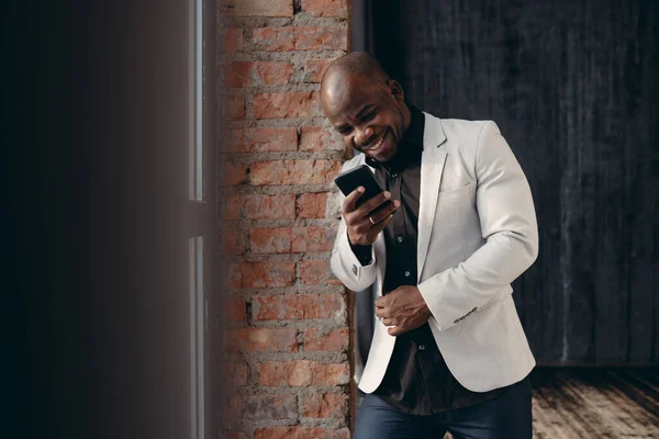 Mężczyzna w beżowej kurtce opierający się o ceglaną ścianę patrząc na telefon i uśmiechając się — Zdjęcie stockowe
