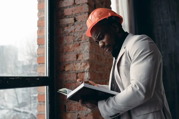 Афроамериканец в строительном шлеме смотрит в окно и пишет в блокноте: — стоковое фото