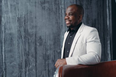 Siyah arka planda koltuğun yanında dururken gülümseyen Afrikalı Amerikalı adam.