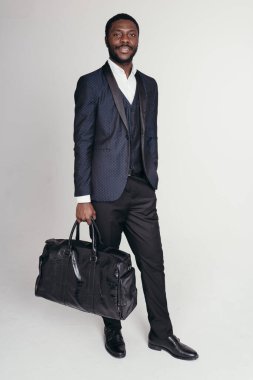 Siyah takım elbiseli Afro-Amerikan bir iş adamı beyaz bir arka planda iş seyahatleri için büyük bir seyahat çantası taşıyor.