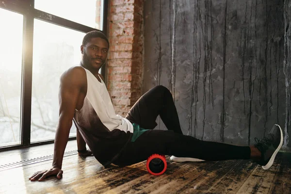 El entrenador de fitness masculino afroamericano resuelve la fascia de la parte posterior del muslo con un rollo de masaje. Se sienta en un estudio loft cerca de una gran ventana al sol —  Fotos de Stock