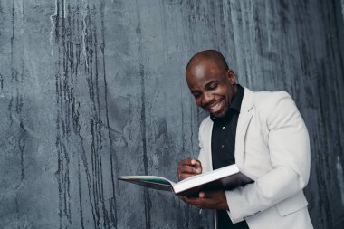 Beyaz ceketli Afro-Amerikan bir adam koyu bir arka planda duruyor bir defter tutuyor ve ona yazıyor.