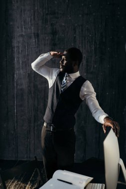 Resmi takım elbiseli yakışıklı adam masanın yanında duruyor, bilgisayarı tutuyor ve yan tarafa bakıyor.