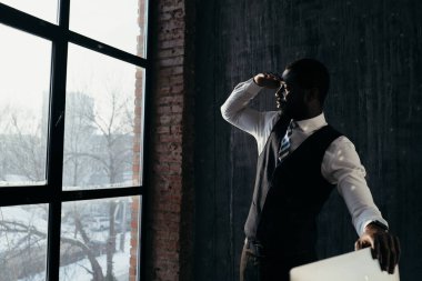 Resmi takım elbiseli yakışıklı adam masanın yanında duruyor, bilgisayarı tutuyor ve yan tarafa bakıyor.