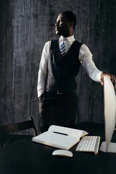 Όμορφος άντρας με επίσημο κοστούμι στέκεται κοντά στο τραπέζι, κρατώντας τον υπολογιστή και κοιτάζει στο πλάι. — Φωτογραφία Αρχείου