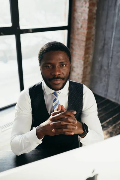 Africano americano homem em uma camisa branca no fundo de uma janela com as palmas das mãos cruzadas olha para a câmera — Fotografia de Stock