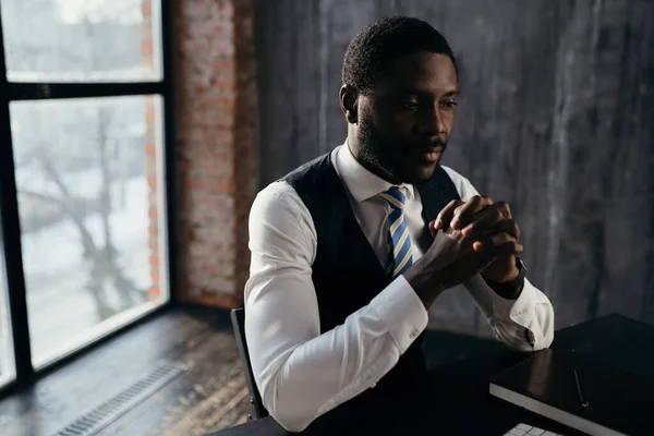 Afro-Amerikaanse man in een wit shirt op de achtergrond van een venster met zijn handpalmen gekruist kijkt naar de camera — Stockfoto