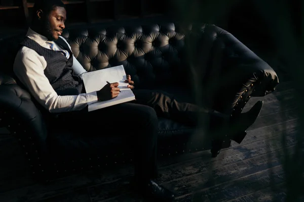 Afroamerykanin siedzi na skórzanej, czarnej sofie, robiąc notatki w notatniku. Koncepcja odpoczynku po pracy — Zdjęcie stockowe