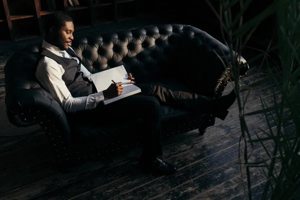 Homem afro-americano senta-se em um sofá preto de couro fazendo anotações em um caderno. Conceito de descanso após o trabalho — Fotografia de Stock
