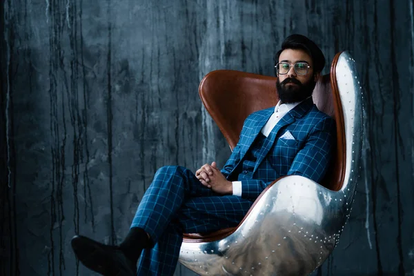 Стильный индус с черной бородой в костюме сидит в кресле и смотрит в камеру — стоковое фото