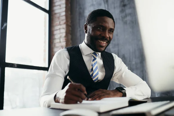 Afroameričan sedí u stolu v kravatě a usmívá se a píše do sešitu — Stock fotografie