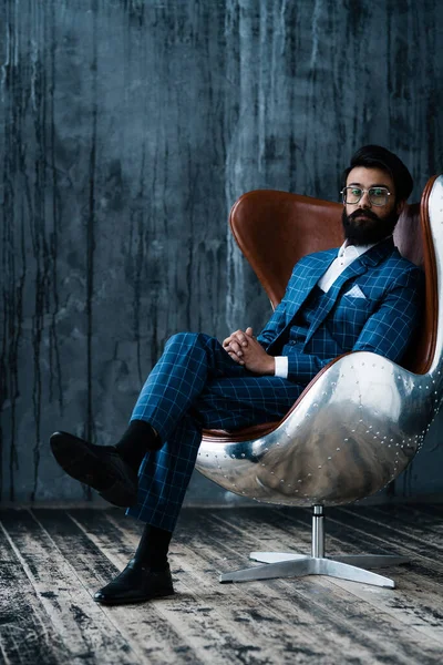 Стильный индус с черной бородой в костюме сидит в кресле и смотрит в камеру — стоковое фото
