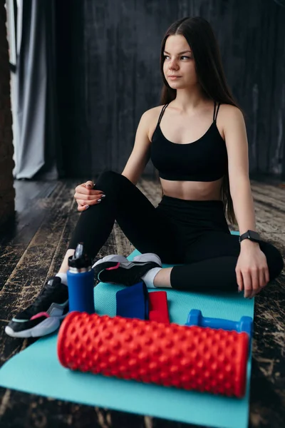 Entrenador en un top negro y polainas se sienta en una alfombra en el gimnasio con un termo y bandas elásticas deportivas — Foto de Stock