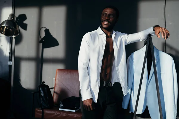 Handsome african american guy stands in a white shirt in an apartment with shadows on the wall leaning on hangers — Stock Photo, Image