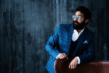 Beyaz gömlekli, siyah sakallı, ciddi bir Hintli arka planda bir sandalyenin yanında duruyor.