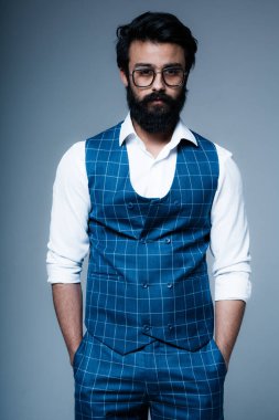 Gözlüklü ve kareli yelek giyen yakışıklı Hintli elleri ceplerinde gri bir arka planda duruyor.