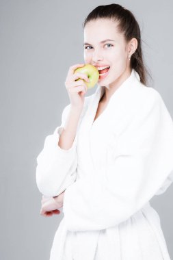 Genç bir kız bir elmayı ısırıyor, gri bir arka planda beyaz bir bornozun içinde duruyor. Diş sağlığı ve uygun beslenme konsepti