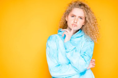 Çekici kızıl saçlı kıvırcık kadın sarı arka planda sessizlik işareti gösteriyor. Mavi kazaklı beyaz kız poz veriyor.