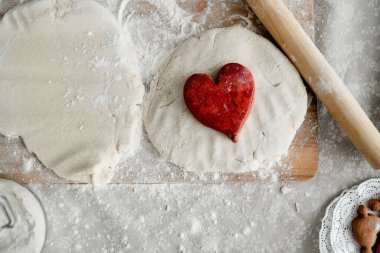 Kırmızı kalp şeklinde bir kurabiye hamurun üzerinde, tahtanın yanında merdanenin yanında.