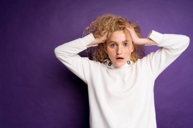 Kafasını iki eliyle tutan bir kadın mor arka planda beyaz bir kazakla saçını mahvediyor.