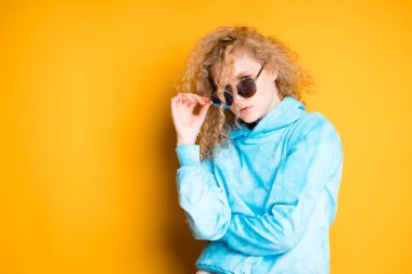 Sarışın, güzel bir kız, parlak bir arka planda dururken eli yüzünde gözlük tutuyor.