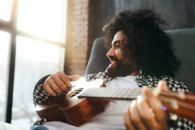 Yakışıklı, siyah saçlı, kanepede uzanmış gitar çalan, gülümseyen ve yan tarafa bakan Mısırlı adam.