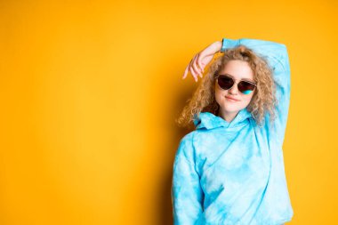 Kıvırcık saçlı güzel bir kız gözlüklü ve bir eli başının arkasında duruyor.