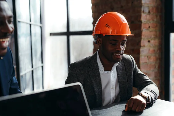 African american man in formal suit sits at a table in a construction helmet next to another worker