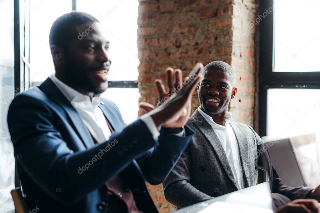 African american man in suit gesturing while talking to partners and sitting next to another smiling worker