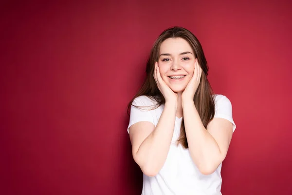 Mujer feliz con frenillos y una camiseta blanca sonríe ampliamente, tocándose las mejillas con las manos —  Fotos de Stock