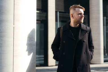 Sırt çantası ve elleri ceplerinde siyah ceketli yakışıklı adam gün ışığında şehirde dolaşıyor.
