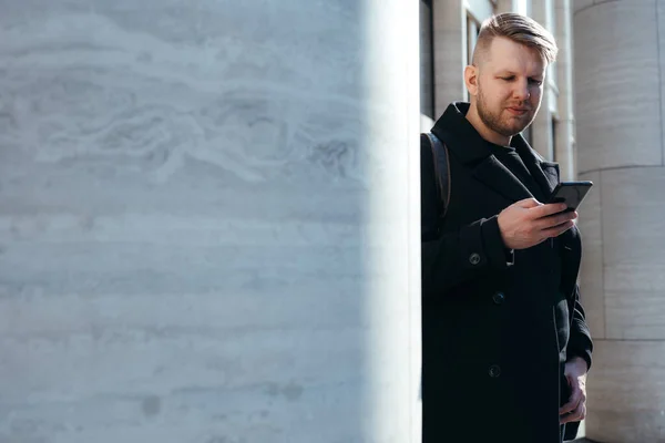 Серьезный человек с бородой держит телефон в руках и работает в нем, стоя среди мраморных колонн — стоковое фото