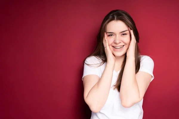 Een mooi meisje in een wit T-shirt met lang haar legt haar handen op haar gezicht en glimlacht breed tussen de beugels — Stockfoto