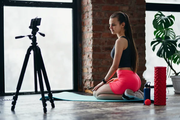 Fitness tränare sitter på mattan registrerar sin träning på en kamera stående på ett stativ i gymmet — Stockfoto