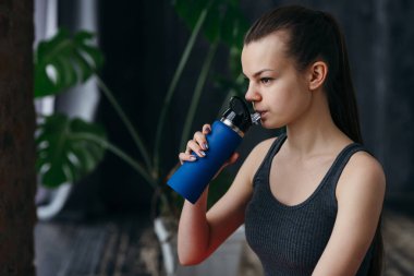 Kuyruklu ve siyah bluzlu güzel bir kız elinde bir termos tutuyor ve spor salonunda egzersiz yaparken termostan içiyor.