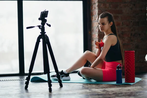 En vacker stretchtränare sitter på en matta i shorat och topp och visar kameran hur en massageboll fungerar på armmusklerna — Stockfoto
