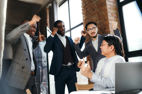 Group of happy asian and black managers rejoice at a successful deal by clenching their hands into fists and raising them up — стоковое фото