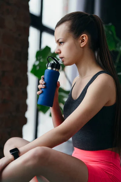 Linda chica en ropa deportiva se sienta de lado en una esterilla de yoga delante de la cámara y bebe agua de una botella mientras la sostiene en su mano —  Fotos de Stock