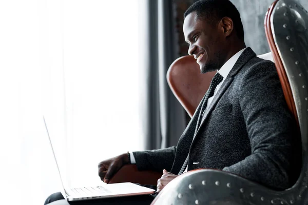 Radosny, ciemnoskóry menadżer siedzi na krześle i pracuje przy laptopie, uśmiechając się szeroko i składając dłoń w miłym geście. Zdjęcie boczne — Zdjęcie stockowe