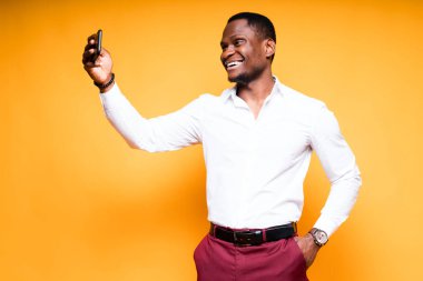Beyaz tişörtlü Afro-Amerikalı adam gülümseyerek selfie çekerken elinde bir telefon tutuyor.