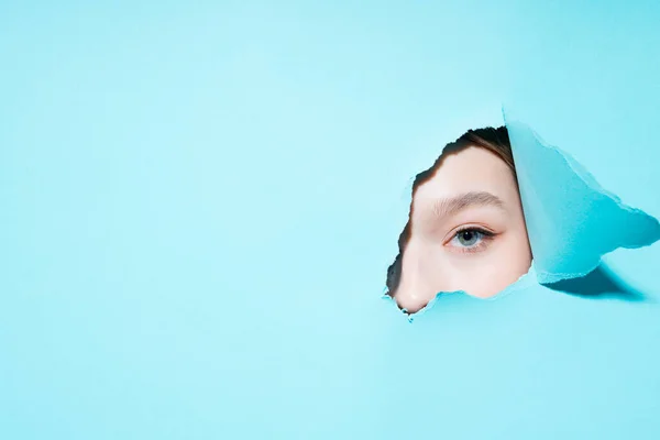 Foto mata seorang gadis yang dicat melihat ke kamera melalui lubang di latar belakang biru — Stok Foto
