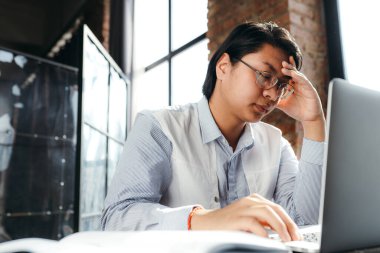 Takım elbiseli Asyalı bir ofis çalışanı dizüstü bilgisayarla uğraşırken şakağına baş ağrısı ile dokunuyor.