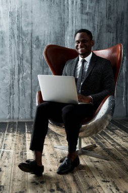 Mutlu Afro-Amerikan ofis çalışanı sandalyeye oturmuş dizüstü bilgisayarla çalışırken gülümsüyor.