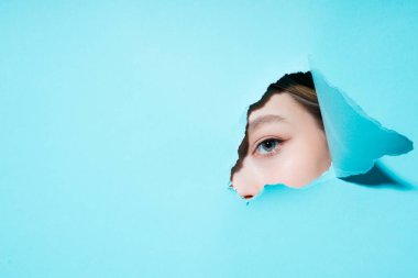 Mavi arka planda bir delikten kameraya bakan genç bir kızın boyalı gözü ve kaşları.