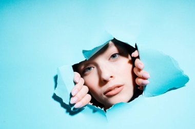 Kızın boyalı yüzü delikten geçerek kameraya doğru gidiyor. Elleriyle yardım ediyor.