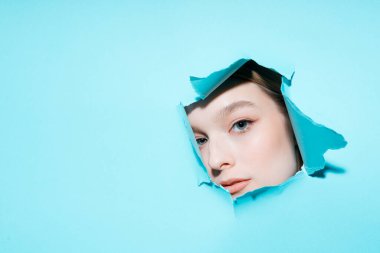 Makyajlı bir kızın yüzü mavi arka plandaki delikten kameraya bakıyor.