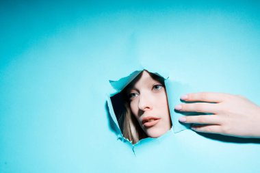 Makyajlı genç bir kız mavi arka plandaki bir delikten yana bakıyor ve elini gösteriyor.